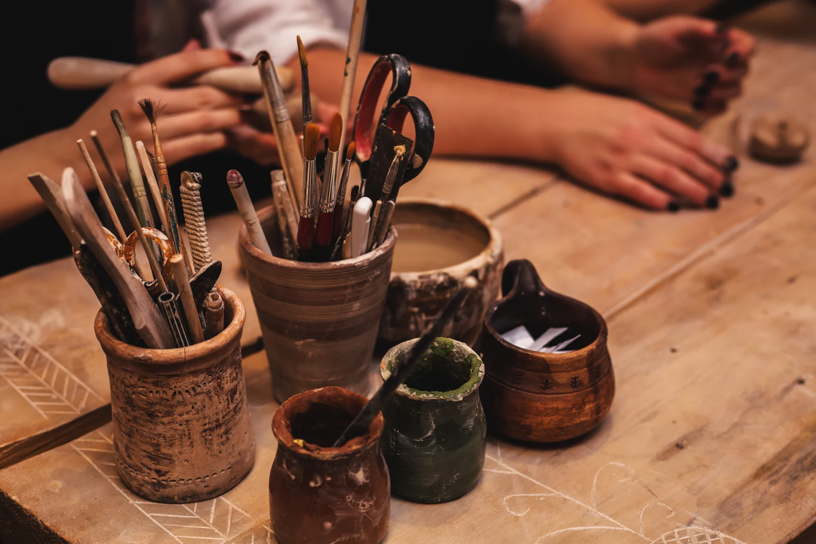 A detailed view of a wooden workbench in the GRAMMA "Atelier," featuring handcrafted clay vessels holding brushes, scissors, and wooden sculpting tools. In the background, the hands of the creators are visible at work.