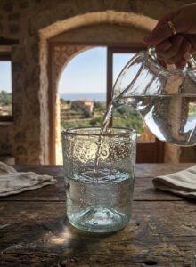 Close-up shot of fresh water from the natural springs of Pagondas in Samos, the headquarters of the House of GRAMMA, being poured into a unique blown glass tumbler with natural dimples. Placed on a rustic wooden table, this authentic piece brings tradition and the refreshing feel of the Greek countryside.