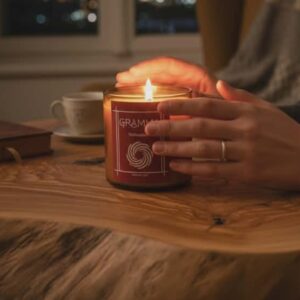 A lit handmade candle featuring the GRAMMA label and labyrinth logo, resting on a natural live-edge wooden table. Two hands gently cup the warm glass. In the softly blurred background, a window with evening lights, a book, and a cup complete a cozy atmosphere of peace and introspection.