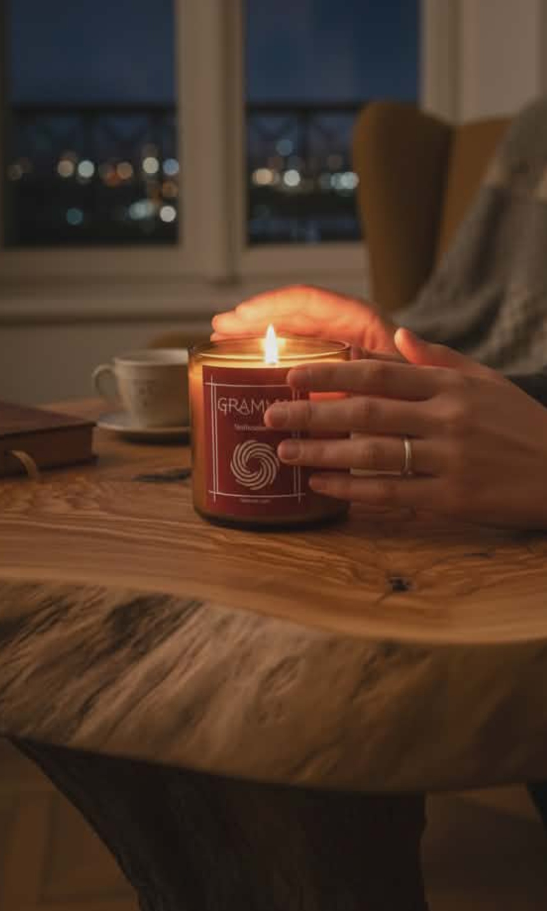 A lit handmade candle featuring the GRAMMA label and labyrinth logo, resting on a natural live-edge wooden table. Two hands gently cup the warm glass. In the softly blurred background, a window with evening lights, a book, and a cup complete a cozy atmosphere of peace and introspection.