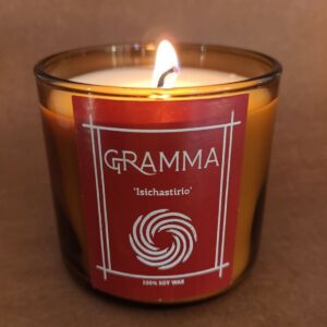 Close-up of a lit, handmade candle made from 100% soy wax. It belongs to the 'Isichastirio' (Sanctuary) chapter of the House of GRAMMA, featuring a distinctive red label with a spiral logo. Placed on an aged, rustic wooden table in warm, low lighting, the candle exudes absolute serenity and coziness.