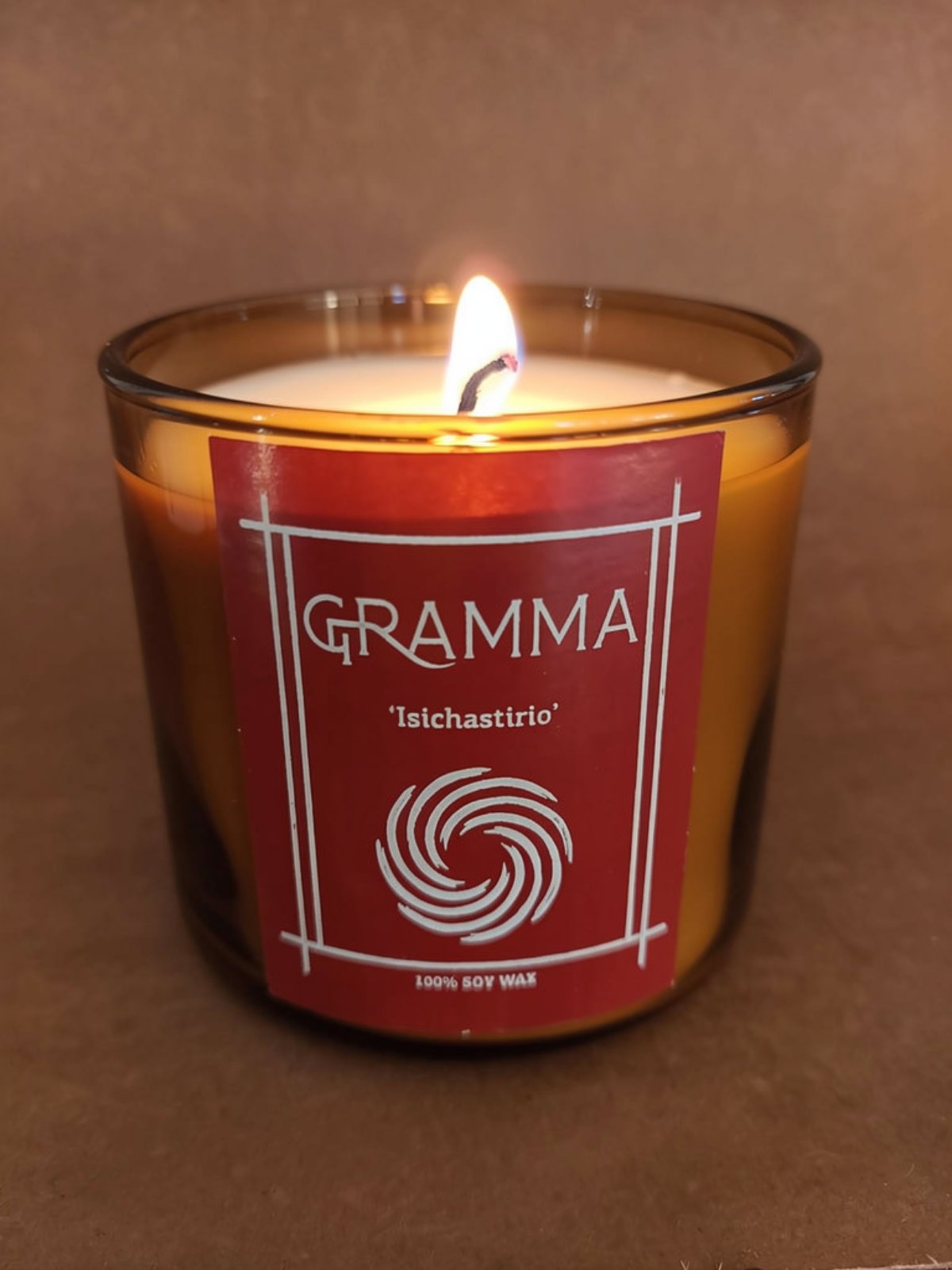 Κοντινή λήψη ενός αναμμένου χειροποίητου κεριού από 100% κερί σόγιας. Ανήκει στο κεφάλαιο 'Ησυχαστήριο' του Οίκου GRAMMA και φέρει χαρακτηριστική κόκκινη ετικέτα με σπειροειδές λογότυπο. Τοποθετημένο πάνω σε ένα παλιό, ρουστίκ ξύλινο τραπέζι σε ζεστό, χαμηλό φωτισμό, το κερί αποπνέει απόλυτη γαλήνη και θαλπωρή.