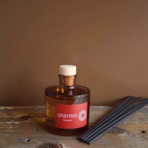 An elegant glass reed diffuser by GRAMMA, resting on an aged wooden surface. The red label features the labyrinth logo. Next to the bottle lie black diffuser sticks, neatly arranged. The background is a warm, earthy brown color, exuding total calmness and the feeling of an Isichastirio