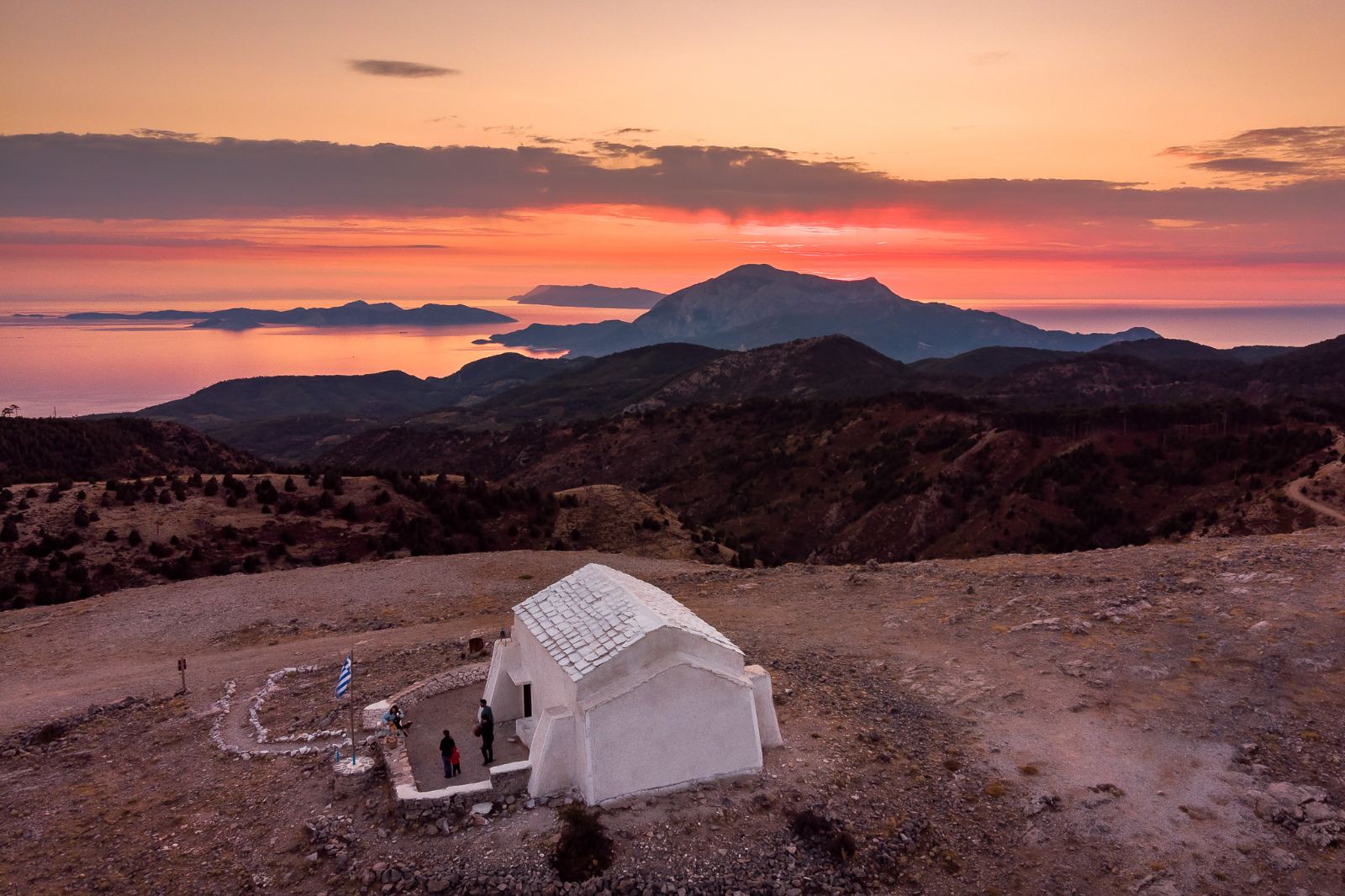 Panoramic view of a traditional white chapel on a mountain peak, overlooking the sea during a breathtaking sunset. This serene landscape embodies the concept of a "Sanctuary" and the deep cultural roots of the House of GRAMMA.