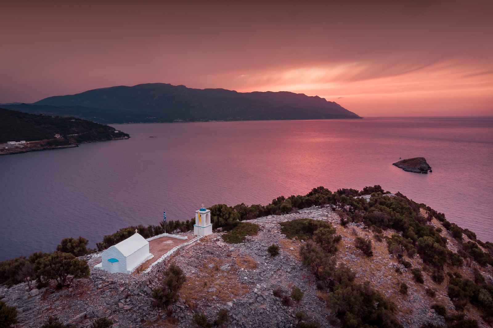 Aerial view of a traditional white chapel on the edge of a rocky cliff, overlooking the sea under the enchanting purple and pink hues of dusk. The landscape exudes absolute serenity, reflecting the concept of the "Sanctuary" and the love for Greek culture championed by the House of GRAMMA.