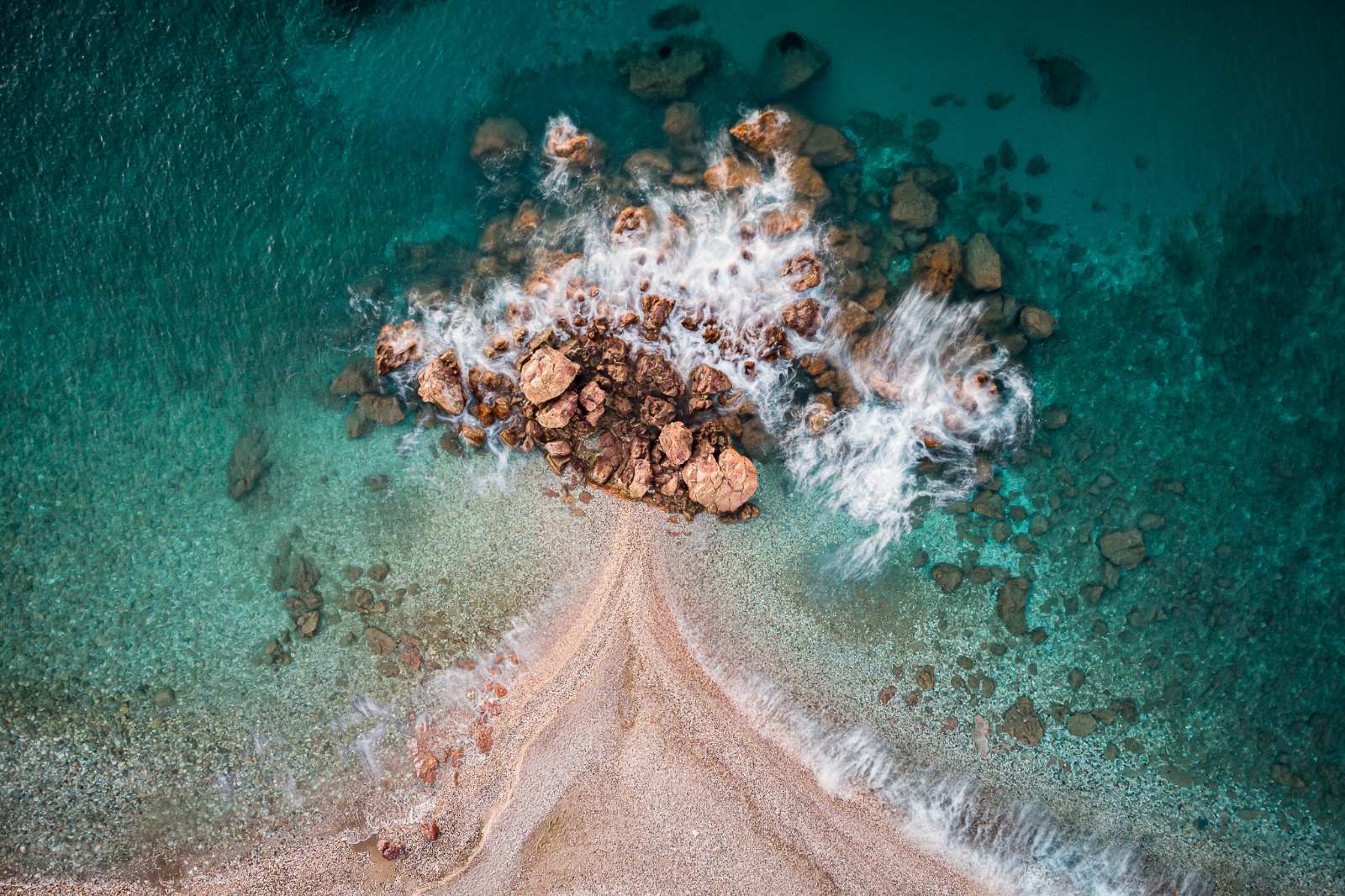 Stunning aerial view where the crystal-clear, turquoise sea waters meet the rugged beauty of the coastal rocks. The harmony of nature's elements is the ultimate source of inspiration for the natural materials and creations of the House of GRAMMA.