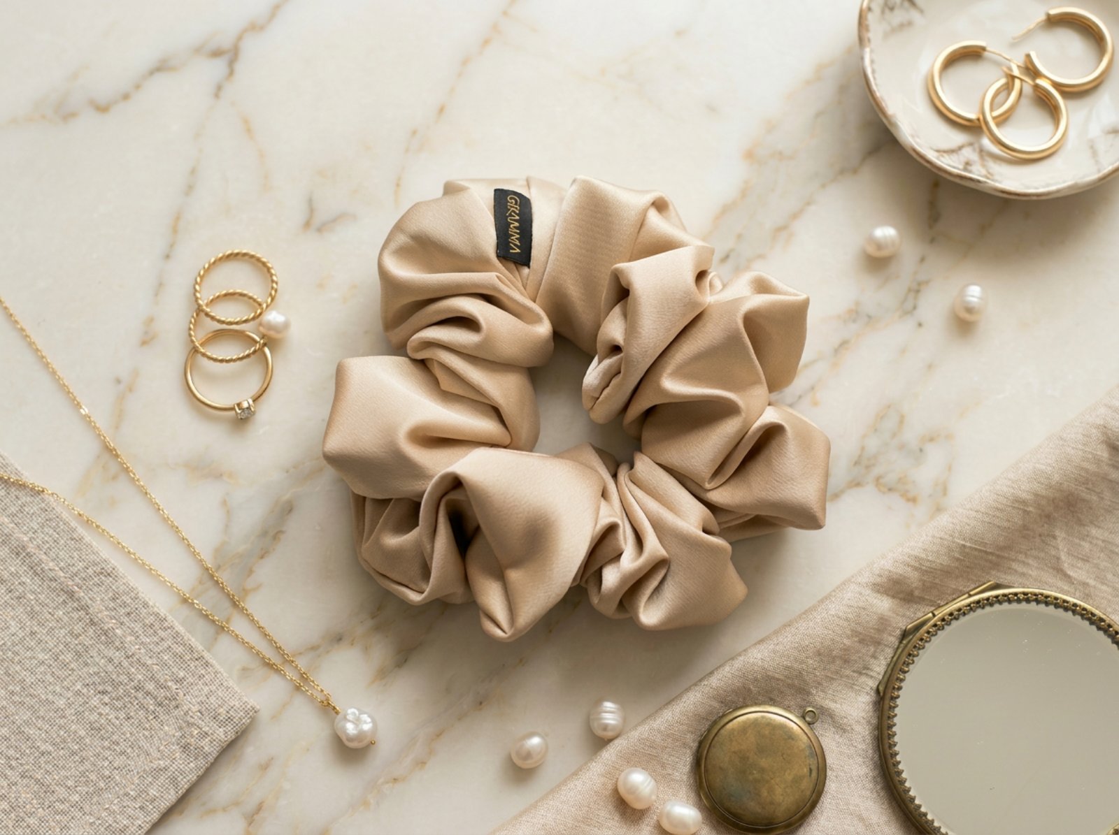 Collection of handmade satin GRAMMA scrunchies in various colors and patterns (Zebra Print, Ochre). Featured in hair styling, close-up with the brand label, and flat lay compositions with jewelry on wooden and marble surfaces.