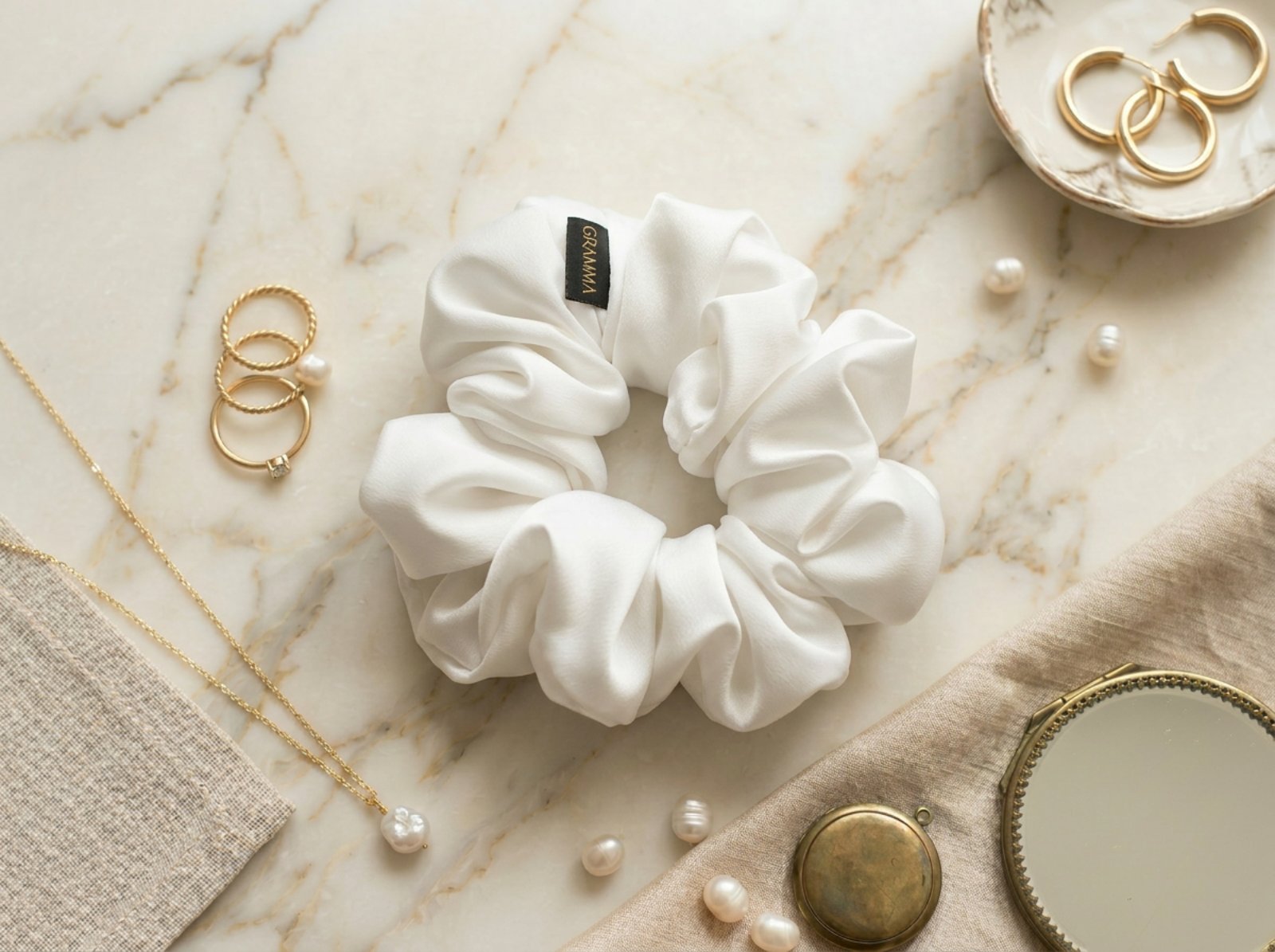 Collection of handmade satin GRAMMA scrunchies in various colors and patterns (Zebra Print, Ochre). Featured in hair styling, close-up with the brand label, and flat lay compositions with jewelry on wooden and marble surfaces.