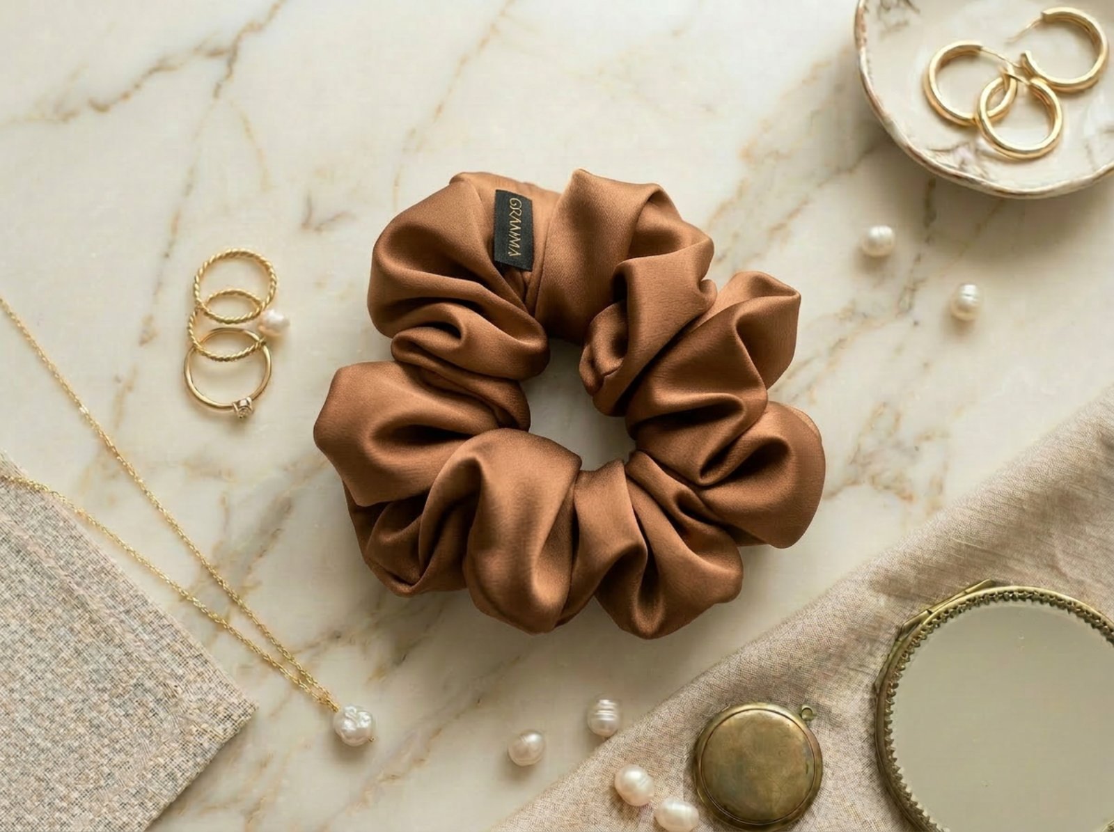Collection of handmade satin GRAMMA scrunchies in various colors and patterns (Zebra Print, Ochre). Featured in hair styling, close-up with the brand label, and flat lay compositions with jewelry on wooden and marble surfaces.