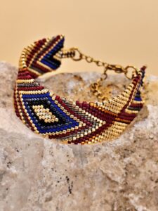 Handcrafted loom-woven cuff bracelet by the House of GRAMMA, featuring Japanese Miyuki beads in a geometric pattern (burgundy, blue, gold). The jewelry rests upon a rough, natural stone.