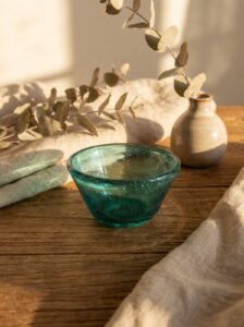 A handmade small bowl made of hand-blown recycled glass in aquamarine tint, placed on a rustic wooden table. The glass features tiny air bubbles, showcasing its artisanal craftsmanship. In the background, a small ceramic bud vase with dried eucalyptus branches and a soft beige linen cloth. The scene is bathed in warm, late afternoon sunlight, creating dramatic and artistic shadows.