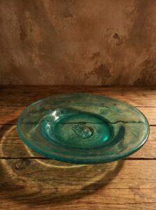 A close-up, atmospheric shot of the handmade small "Limni" plate made from hand-blown recycled glass. The plate features a deep aquamarine color and is placed on a rustic wooden surface. In its center, the embossed seal of authenticity with the GRAMMA emblem is clearly visible. Natural sunlight creates beautiful reflections, highlighting the wavy texture of the glass.