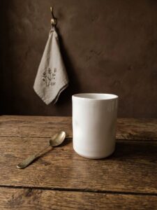 Large handmade white ceramic glass by GRAMMA next to a brass spoon, resting on a rustic wooden table with a linen towel in the background.