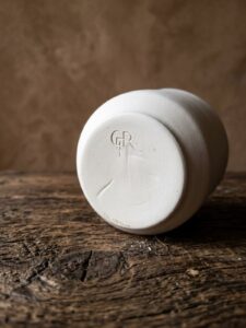 Close-up of the embossed GRAMMA brand stamp on the base of white handmade ceramics, resting on a rustic wooden surface.