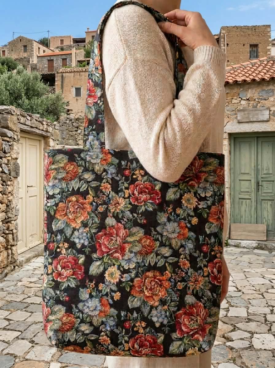 Floral shoulder bag on a dark background, carried over the shoulder of a person in a cream knit, in front of stone houses